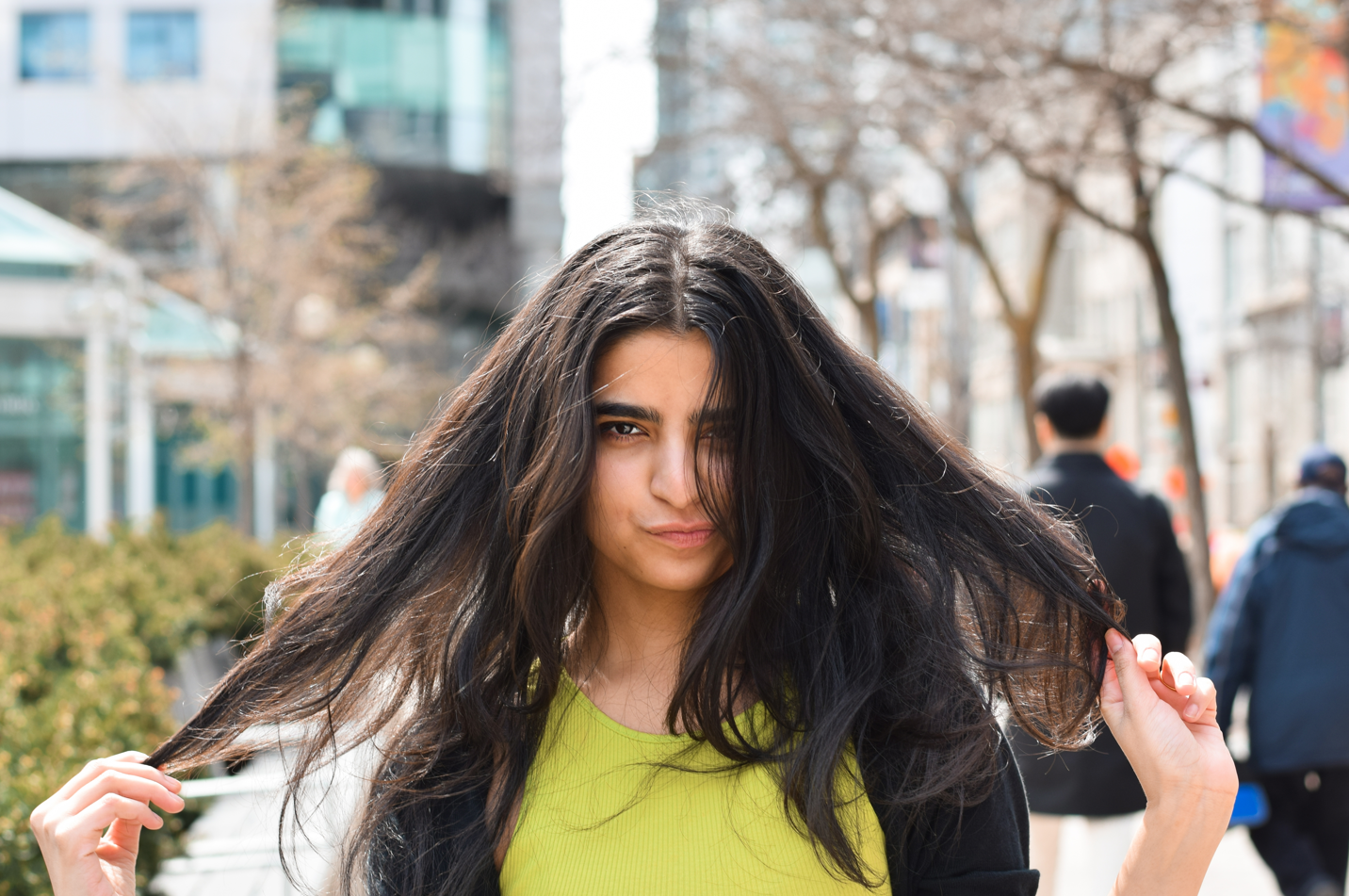 A young girl pulls at her frizzy hair while standing outside in a crowded public place. Concept of how to reduce frizz, tame frizzy hair using hair building fibers.